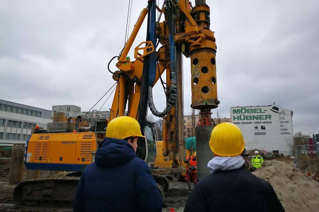 Zwei Personen mit gelben Schutzhelmen beobachten ein großes Bohrgerät auf einer Baustelle.