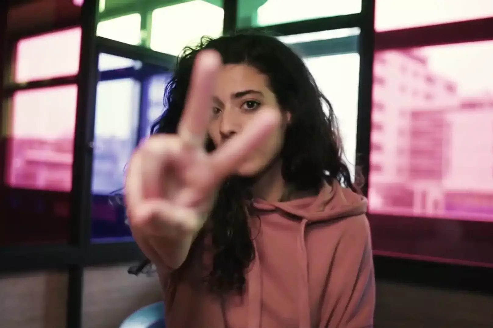 Frau in einem rosafarbenen Hoodie zeigt ein Victory-Zeichen in die Kamera und schaut zwischen Zeige- und Mittelfinger hindurch.
