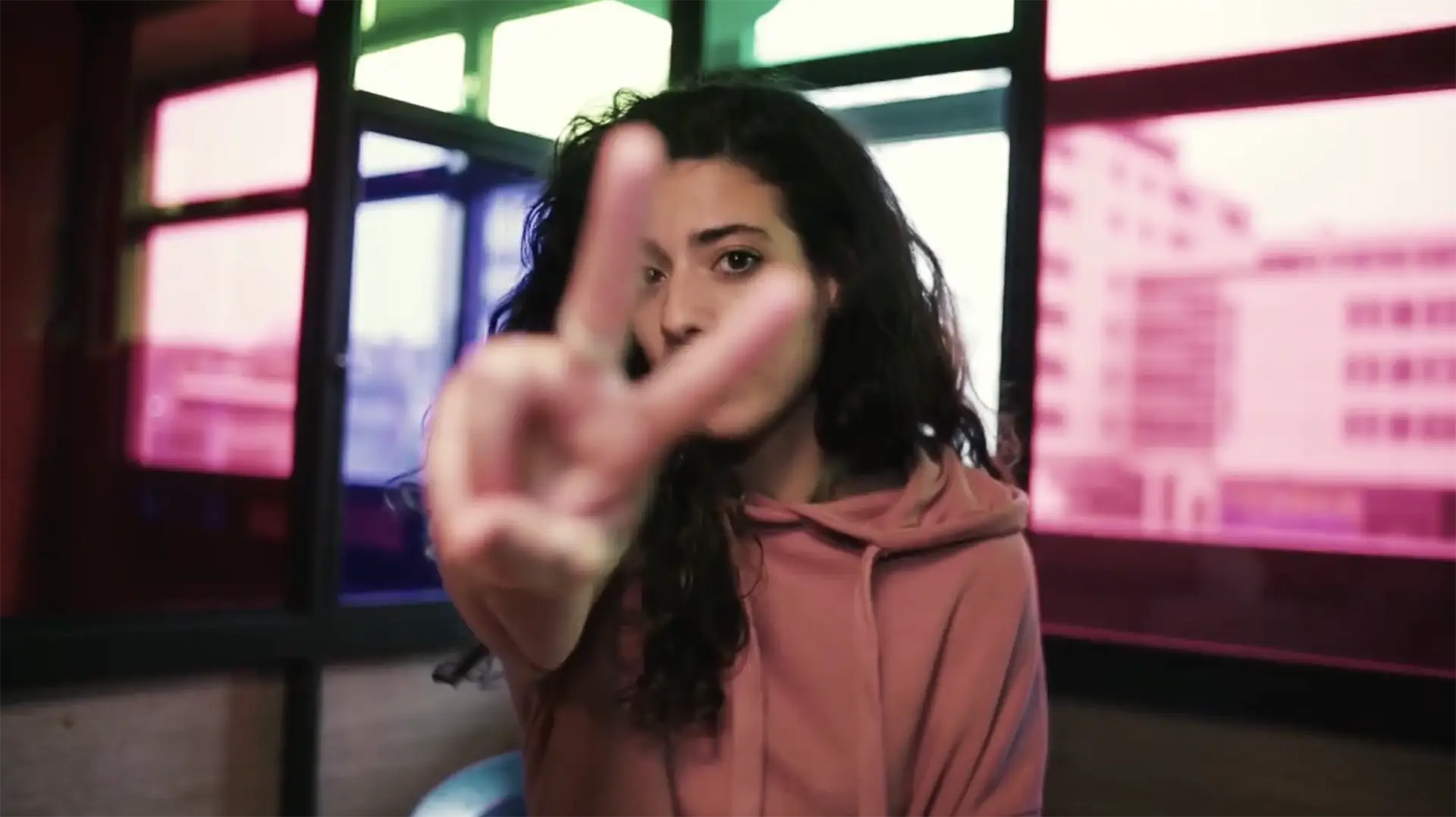 Frau in einem rosafarbenen Hoodie zeigt ein Victory-Zeichen in die Kamera und schaut zwischen Zeige- und Mittelfinger hindurch.