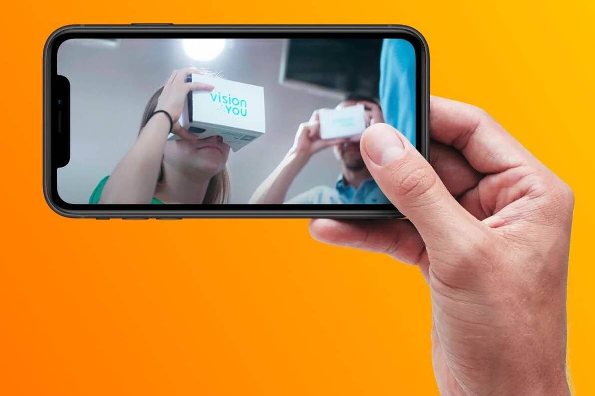 Hand hält ein Smartphone mit einem Foto von zwei Personen, die VR-Brillen von visionYOU testen.