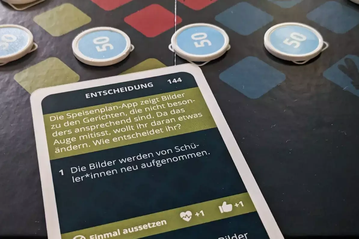 Spielplan von „Vision Mensa“ mit Spielfiguren, Spielgeld und einer Entscheidungskarte zur Gestaltung einer Speiseplan-App.