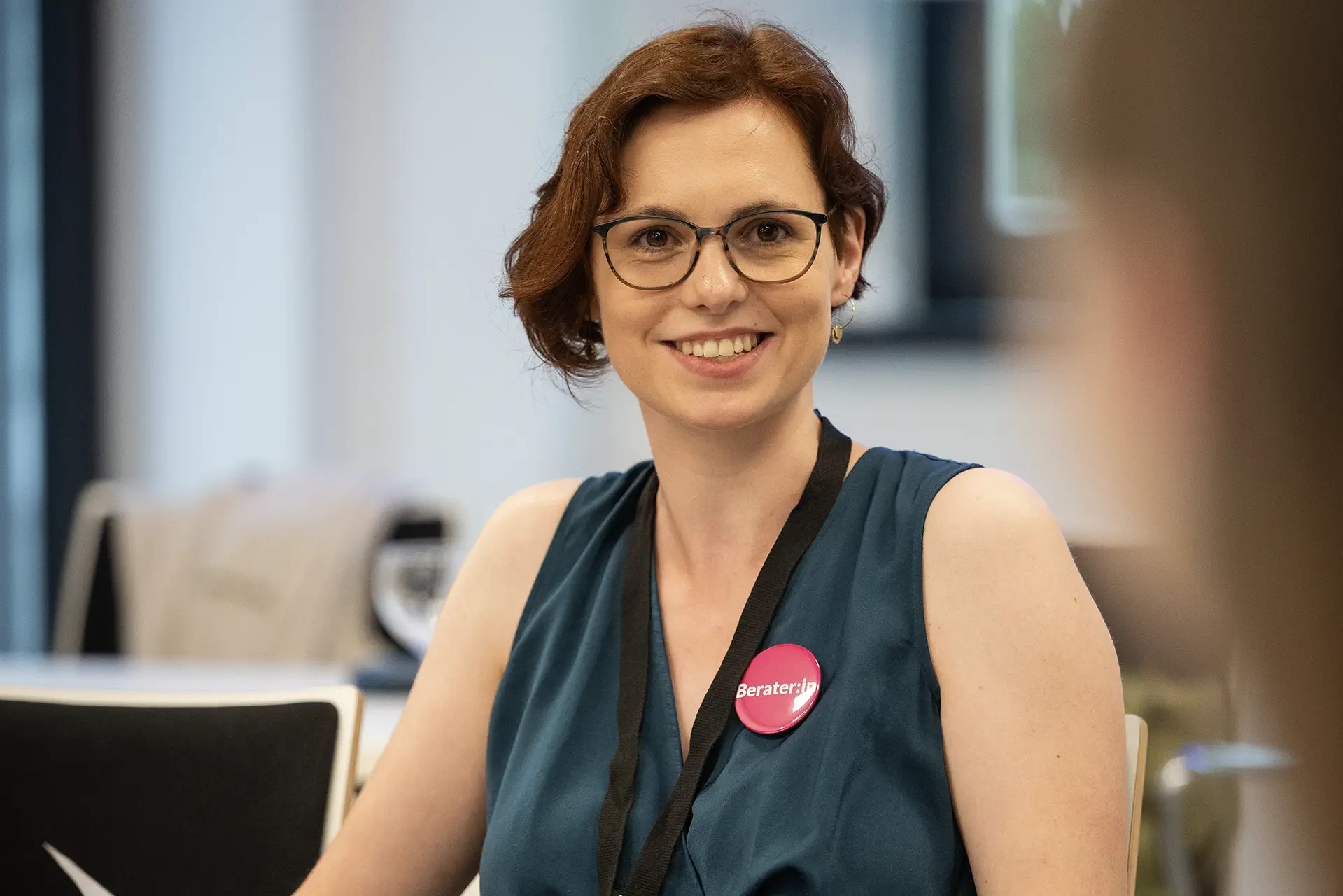 Lächelnde Beraterin mit Brille und pinkfarbenem „Berater:in“-Button bei einer Veranstaltung.